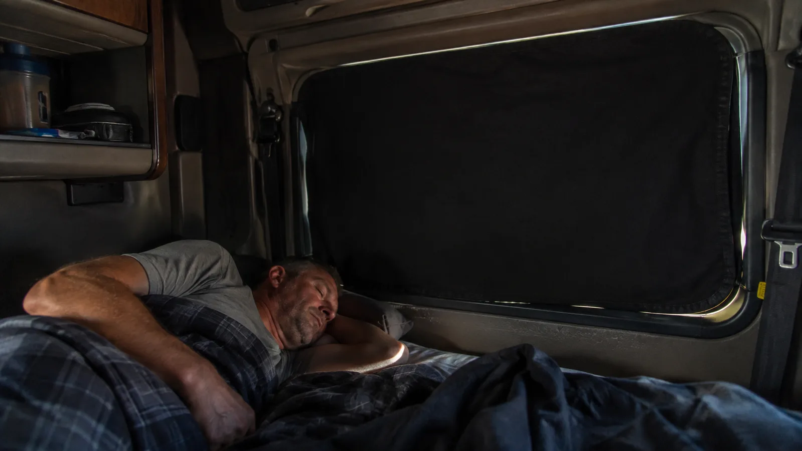A dark truck sleeper cab window fully covered by a black portable blackout shade, cab interior dim and restful at midday.