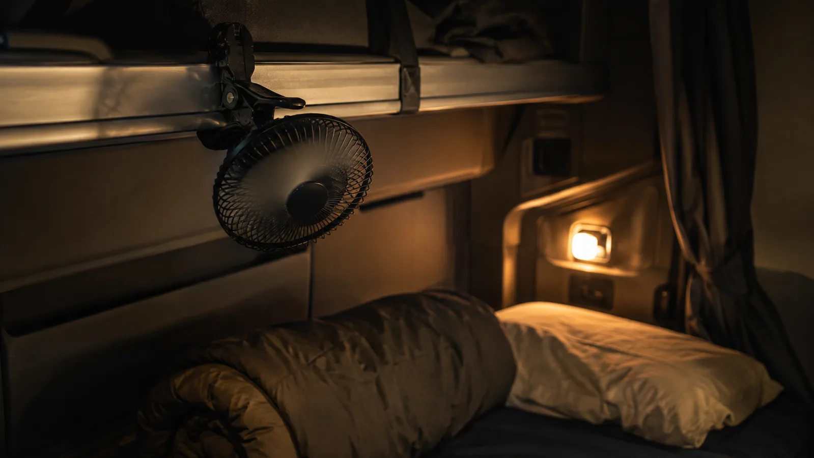 A small clip-on fan clamped to a sleeper cab bunk rail at night, blades in motion