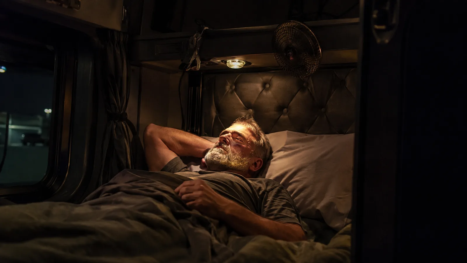 A small clip-on fan clamped to a bunk rail inside a truck sleeper cab at night, warm indigo cab lighting