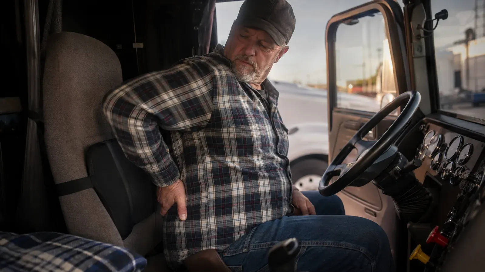 Truck driver adjusting a black lumbar support pillow strapped to his seat inside a semi truck cab