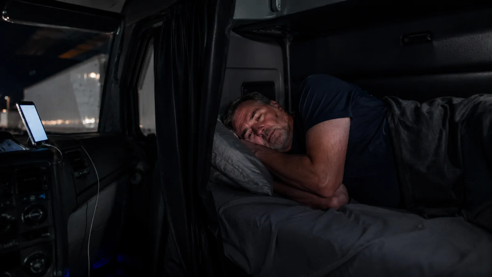 A truck driver lying in a sleeper cab bunk at night with small sleep earbuds in his ears, dashboard lights glowing faintly in the background