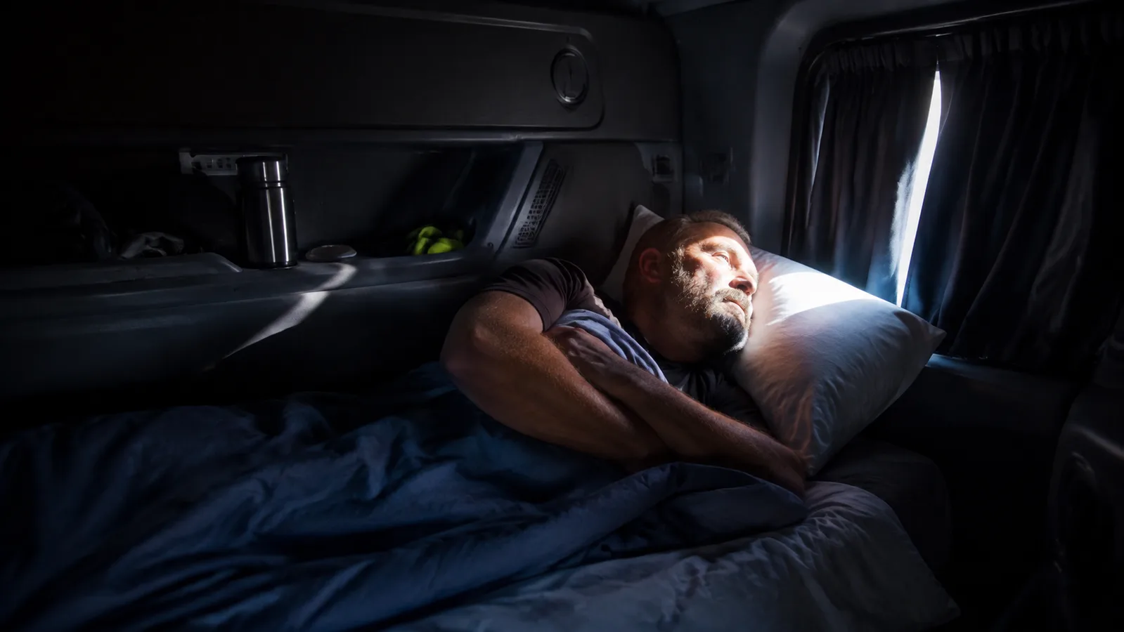 A dark truck cab bunk lit up by a harsh security light shining through the windshield curtain