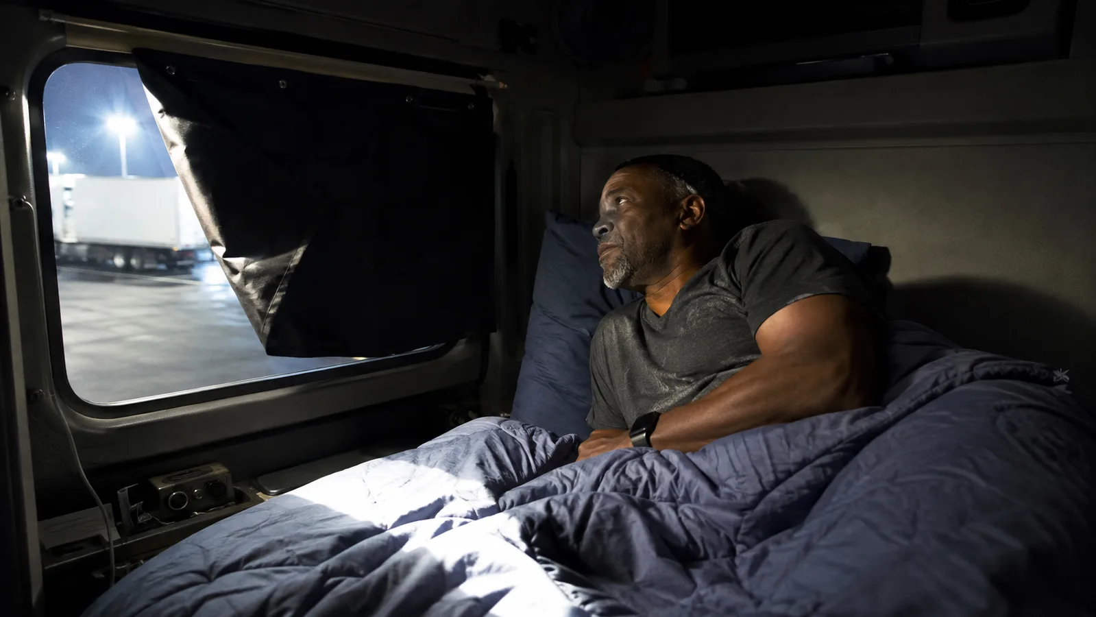 A dim truck sleeper cab at dawn with a strip of harsh parking lot light cutting across the bunk where a blackout shade has sagged at one corner